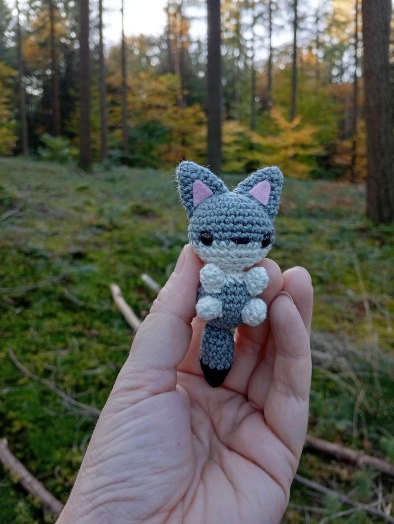 May include: A handmade crochet fox figurine in shades of gray, white, and black, with pink inner ears. The small, detailed fox is held in a hand against a blurred forest background, showcasing the craft's intricate details.