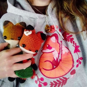 May include: Two crocheted fox plushies in orange and brown with green and brown clothing, sitting on a gray hoodie with a red embroidered fox design and a brown and orange gemstone necklace.