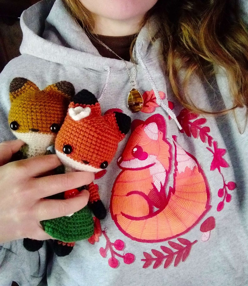 May include: Two crocheted fox plushies in orange and brown with green and brown clothing, sitting on a gray hoodie with a red embroidered fox design and a brown and orange gemstone necklace.