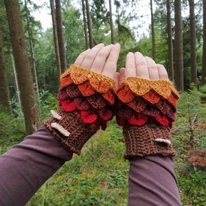 Puede incluir: Guantes sin dedos de ganchillo en tonos marr&oacute;n, rojo, naranja y amarillo, que se asemejan a escamas de drag&oacute;n. Los guantes tienen un dise&ntilde;o texturizado y est&aacute;n acentuados con peque&ntilde;os adornos de color claro. El fondo es una escena de bosque.