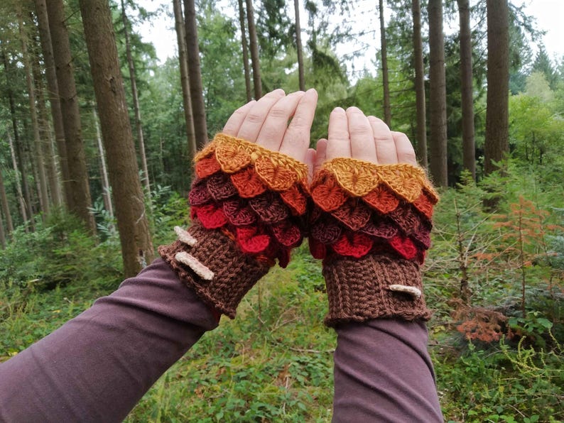 Puede incluir: Guantes sin dedos de ganchillo en tonos marr&oacute;n, rojo, naranja y amarillo, que se asemejan a escamas de drag&oacute;n. Los guantes tienen un dise&ntilde;o texturizado y est&aacute;n acentuados con peque&ntilde;os adornos de color claro. El fondo es una escena de bosque.