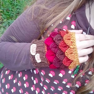 Puede incluir: Calentadores de brazo de ganchillo en tonos marr&oacute;n, rojo y naranja. Los calentadores de brazo presentan un dise&ntilde;o de escamas, con un pu&ntilde;o marr&oacute;n adornado con acentos blancos en forma de hongo. La manga es visible en color marr&oacute;n oscuro.