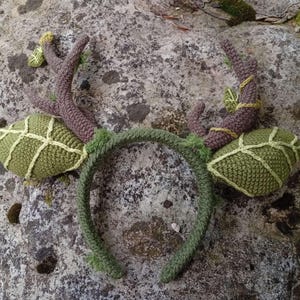May include: A handmade, green crochet headband with attached antlers and leaf-shaped accents. The antlers are brown with yellow accents, and the leaves are a lighter green with a grid pattern. The headband is a darker green, creating a whimsical, nature-inspired accessory.