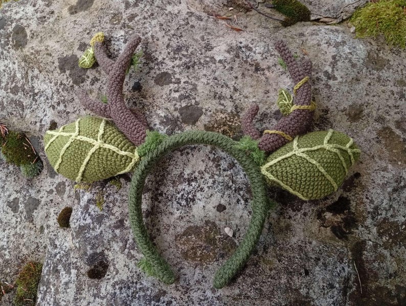 May include: A handmade, green crochet headband with attached antlers and leaf-shaped accents. The antlers are brown with yellow accents, and the leaves are a lighter green with a grid pattern. The headband is a darker green, creating a whimsical, nature-inspired accessory.
