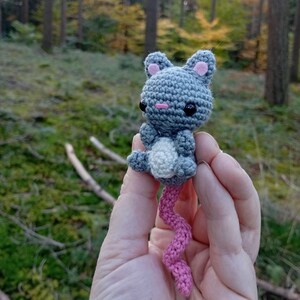 May include: A handmade crochet mouse toy in gray, white, and pink. The mouse has black eyes, pink inner ears, and a long, curly pink tail. The toy is held against a blurred forest background.