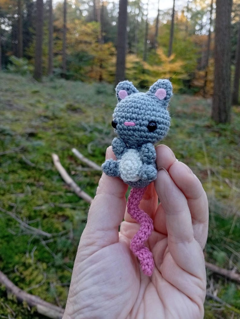 May include: A handmade crochet mouse toy in gray, white, and pink. The mouse has black eyes, pink inner ears, and a long, curly pink tail. The toy is held against a blurred forest background.