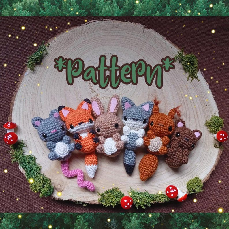 May include: A collection of handmade crochet woodland animal figures, including a gray mouse, orange fox, brown bunny, gray fox, orange squirrel, and brown bear. The word "Pattern" is written in green script on a wooden slice, surrounded by moss and red mushroom decorations.