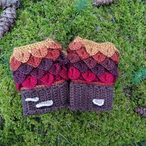 Puede incluir: Un par de guantes sin dedos de ganchillo hechos a mano con un dise&ntilde;o de escamas de drag&oacute;n. Los guantes presentan un degradado de color de oro a rojo y morado. Los pu&ntilde;os son marrones con detalles blancos, sobre un fondo de musgo verde.