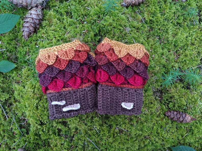Puede incluir: Un par de guantes sin dedos de ganchillo hechos a mano con un dise&ntilde;o de escamas de drag&oacute;n. Los guantes presentan un degradado de color de oro a rojo y morado. Los pu&ntilde;os son marrones con detalles blancos, sobre un fondo de musgo verde.