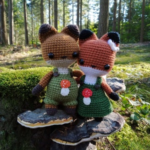 May include: Two crocheted fox plushies wearing overalls and mushroom patches, sitting on a layered mushroom-shaped stump in a forest setting.
