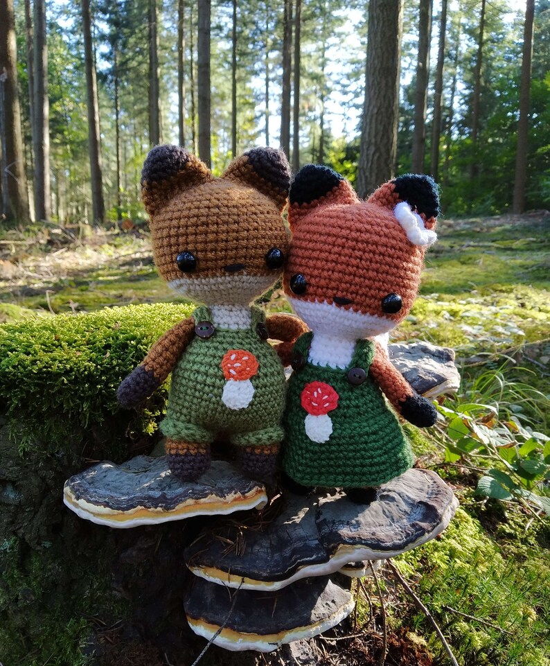 May include: Two crocheted fox plushies wearing overalls and mushroom patches, sitting on a layered mushroom-shaped stump in a forest setting.