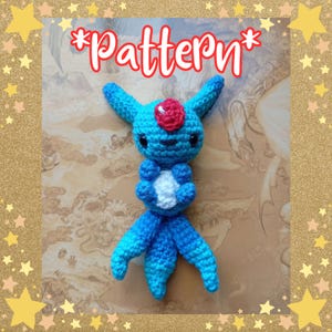 May include: A handmade crochet creature in shades of blue, with a red accent on its head and a white chest. The word "Pattern" is written in red above the creature. The creature has large, black eyes and a whimsical design.