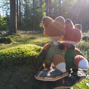 May include: Two crocheted fox plushies, one brown and one green, are sitting on a mushroom-shaped rock in a forest setting. The plushies are facing the same direction, with the brown fox in front of the green fox.