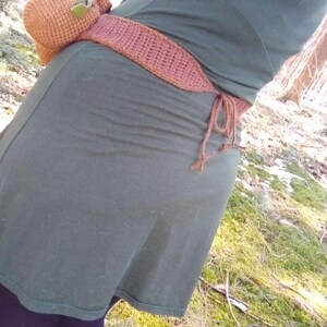 K&ouml;nnte beinhalten: Eine braune geh&auml;kelte Tasche mit einem gr&uuml;nen Blatt daran, die als G&uuml;rtel um die Taille einer Person getragen wird. Die Tasche ist an einem braunen geh&auml;kelten G&uuml;rtel mit einer Schleifenverschlusses befestigt.