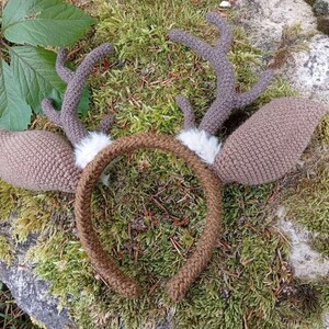 May include: A handmade, brown crocheted deer antler headband with matching ears and a brown band. The antlers are a darker brown and have a textured appearance. White faux fur accents the base of the antlers. The headband is set against a mossy background.