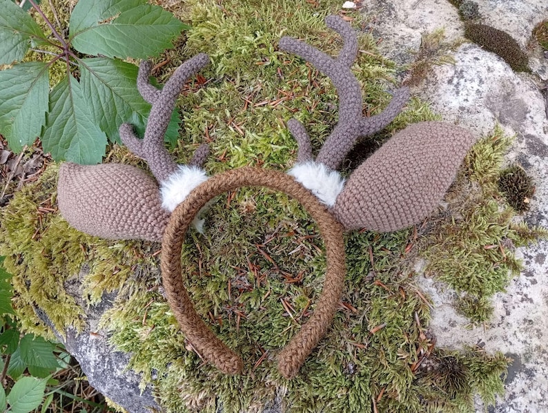 May include: A handmade, brown crocheted deer antler headband with matching ears and a brown band. The antlers are a darker brown and have a textured appearance. White faux fur accents the base of the antlers. The headband is set against a mossy background.
