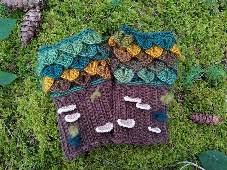 Puede incluir: Un par de guantes sin dedos hechos a mano con ganchillo. Los guantes presentan una base marr&oacute;n con detalles de setas blancas y una secci&oacute;n superior con detalles en forma de hojas verdes, turquesas, marrones y amarillas. Sobre un fondo de musgo.