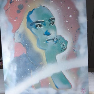 Può includere: Un'opera d'arte rettangolare realizzata con vernice spray su una superficie piana che presenta un ritratto stilizzato di una persona con tonalità di pelle blu e verde acqua. Lo sfondo è composto da una nebbia rosa e bianca con schizzi di vernice.