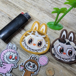 May include: Four embroidered patches featuring cartoon characters with rabbit ears and sharp teeth. One patch is pink, one is brown holding a pink balloon and a cup, one is yellow with brown spots, and one is brown with dark spots. A black lighter is nearby.