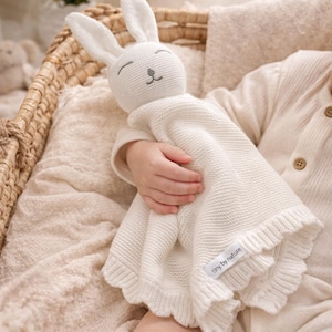 May include: A white knitted bunny security blanket with a ruffled edge. The bunny has long ears and a stitched face. The blanket is held by a child. The label reads "tiny by nature".