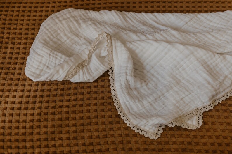 May include: A white, crinkled muslin blanket with a delicate lace trim along the edges. The blanket is folded and draped, showcasing its soft texture. The background is a textured, brown surface.