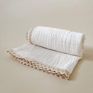 May include: A rolled white blanket with a beige crocheted trim. The blanket is made of a soft, textured fabric and is partially unrolled, revealing the crocheted edge. The neutral color palette makes it suitable for various home decor styles.