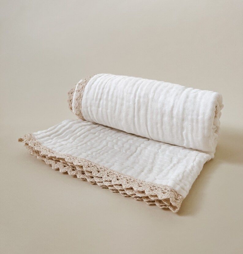 May include: A rolled white blanket with a beige crocheted trim. The blanket is made of a soft, textured fabric and is partially unrolled, revealing the crocheted edge. The neutral color palette makes it suitable for various home decor styles.