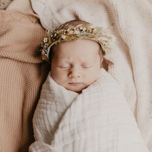 May include: A newborn baby wrapped in a white muslin blanket, wearing a delicate floral crown. The baby is lying on a soft, textured blanket. The color palette is soft and neutral, with shades of beige and cream.