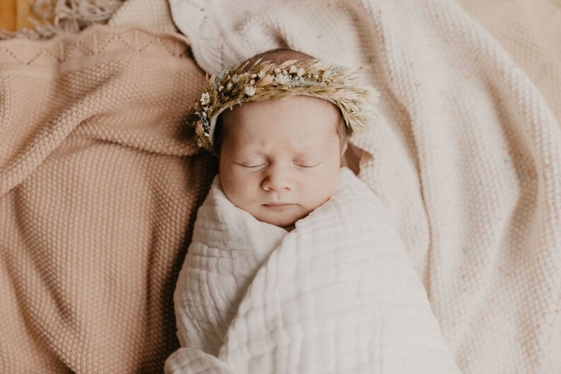 May include: A newborn baby wrapped in a white muslin blanket, wearing a delicate floral crown. The baby is lying on a soft, textured blanket. The color palette is soft and neutral, with shades of beige and cream.