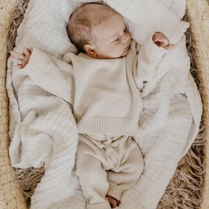 May include: A newborn baby wearing a beige knit sweater and pants set, lying in a woven basket lined with a white muslin blanket. The baby is nestled on a soft, textured surface, creating a cozy and comfortable scene.