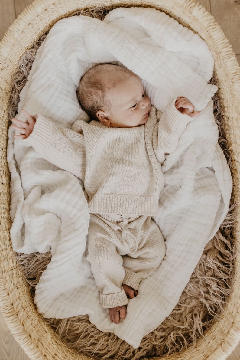 May include: A newborn baby wearing a beige knit sweater and pants set, lying in a woven basket lined with a white muslin blanket. The baby is nestled on a soft, textured surface, creating a cozy and comfortable scene.