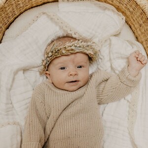 May include: A baby wearing a beige sweater and a floral crown lies in a woven basket. The baby is on a white blanket with lace trim. The basket is made of woven straw.