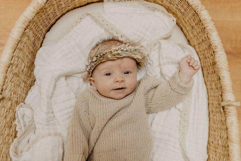 May include: A baby wearing a beige sweater and a floral crown lies in a woven basket. The baby is on a white blanket with lace trim. The basket is made of woven straw.