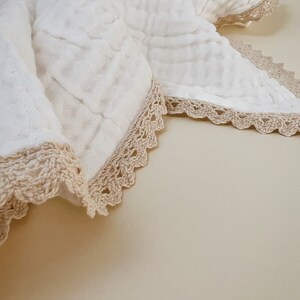 May include: A white, textured blanket with a crocheted, beige lace trim. The blanket is folded, revealing the delicate texture of the fabric and the intricate detail of the lace edging. The overall aesthetic is soft and inviting.