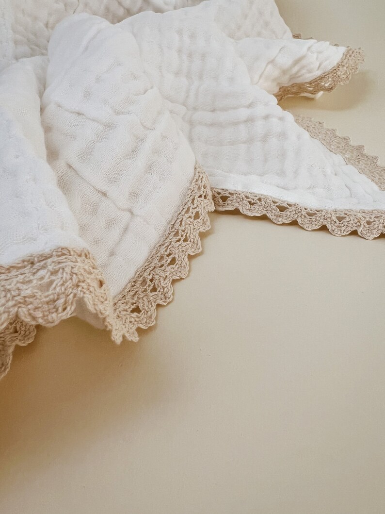 May include: A white, textured blanket with a crocheted, beige lace trim. The blanket is folded, revealing the delicate texture of the fabric and the intricate detail of the lace edging. The overall aesthetic is soft and inviting.