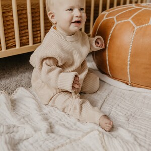 May include: A baby wearing a beige knit sweater and matching pants sits on a white blanket. The baby is near a wooden crib and a brown leather ottoman. The outfit is a neutral color, suitable for a baby.