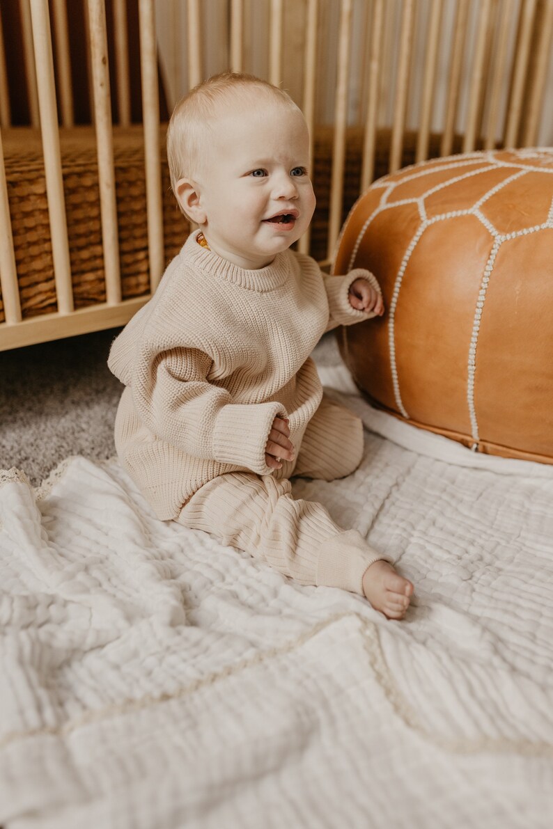 May include: A baby wearing a beige knit sweater and matching pants sits on a white blanket. The baby is near a wooden crib and a brown leather ottoman. The outfit is a neutral color, suitable for a baby.