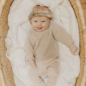 May include: A smiling baby wearing a beige sweater, pants, and a floral crown, lying in a woven basket lined with a white blanket. The baby is sticking out their tongue. The basket is oval-shaped and has a natural, light brown color.