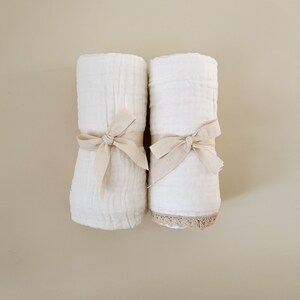 May include: Two rolled white fabric items, possibly towels or blankets, are tied with beige ribbons. One item has a beige lace trim. The items are set against a neutral background, creating a soft, minimalist aesthetic.