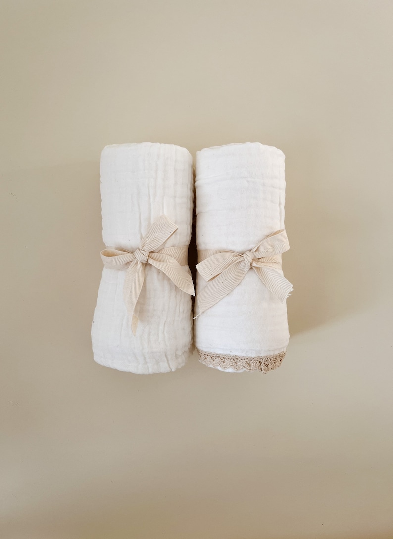 May include: Two rolled white fabric items, possibly towels or blankets, are tied with beige ribbons. One item has a beige lace trim. The items are set against a neutral background, creating a soft, minimalist aesthetic.