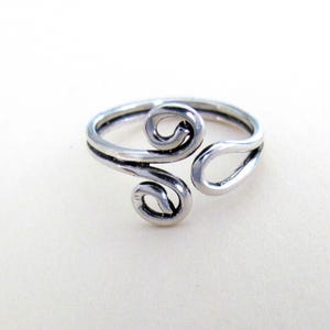 May include: A silver-colored toe ring with a unique design. The ring features two swirling, scroll-like shapes and a teardrop-shaped opening. The ring is made of a shiny, metallic material and is set against a white background.