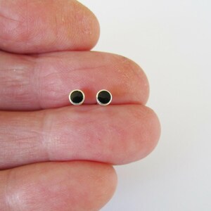 May include: Two small, round black stud earrings with silver-colored settings. The earrings are displayed on a light background.