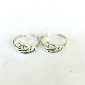 May include: Two silver-colored toe rings. Each ring features a wave-like design in the center. The rings are adjustable and have an open design. The rings are on a white surface.