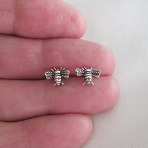 May include: A pair of small, silver-colored bee stud earrings. Each earring features a detailed bee design with wings and body segments. The earrings are displayed against a neutral background.