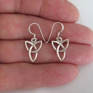 May include: A pair of silver Celtic knot earrings with a triangular shape. The earrings are on a hand.