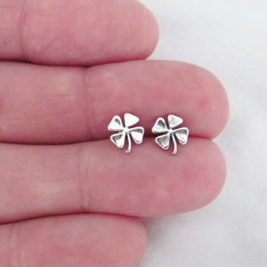May include: A pair of small, silver-colored clover-shaped stud earrings. Each earring features four detailed leaves, showcasing a classic lucky charm design. The earrings are displayed against a neutral background.
