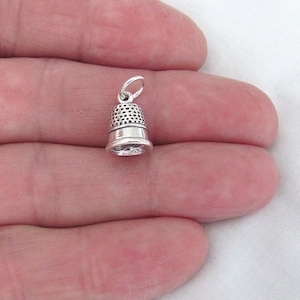 May include: Silver thimble charm with a textured surface. The charm has a loop at the top for attaching to a necklace or bracelet.
