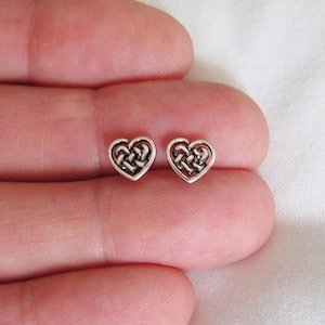 May include: A pair of small, silver-colored heart-shaped stud earrings. Each earring features a Celtic knot design within the heart's outline. The earrings are displayed against a soft, off-white background.
