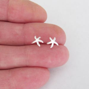 May include: A pair of small, silver starfish stud earrings. The earrings are simple in design, with five arms extending from a central point. The earrings are displayed on a light background.