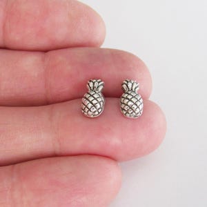 May include: A pair of silver-colored pineapple stud earrings. Each earring features a detailed pineapple design with a textured surface. The earrings are small and delicate, perfect for everyday wear.
